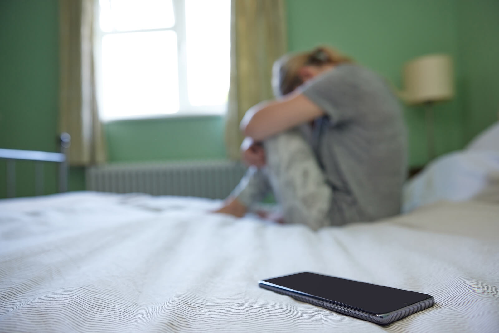Young woman sits on the bed with her phone out of reeach. Avoiding life is not the path to recovery. Get help with online PTSD therapy in Ohio and Kentucky.