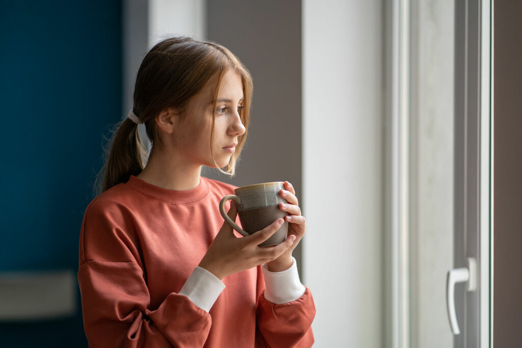 A young female sexual assault survivor looks out the window while drinking a cup of coffee representing an individual who could benefit from Online Therapy for Sexual Assault Survivors in Ohio.