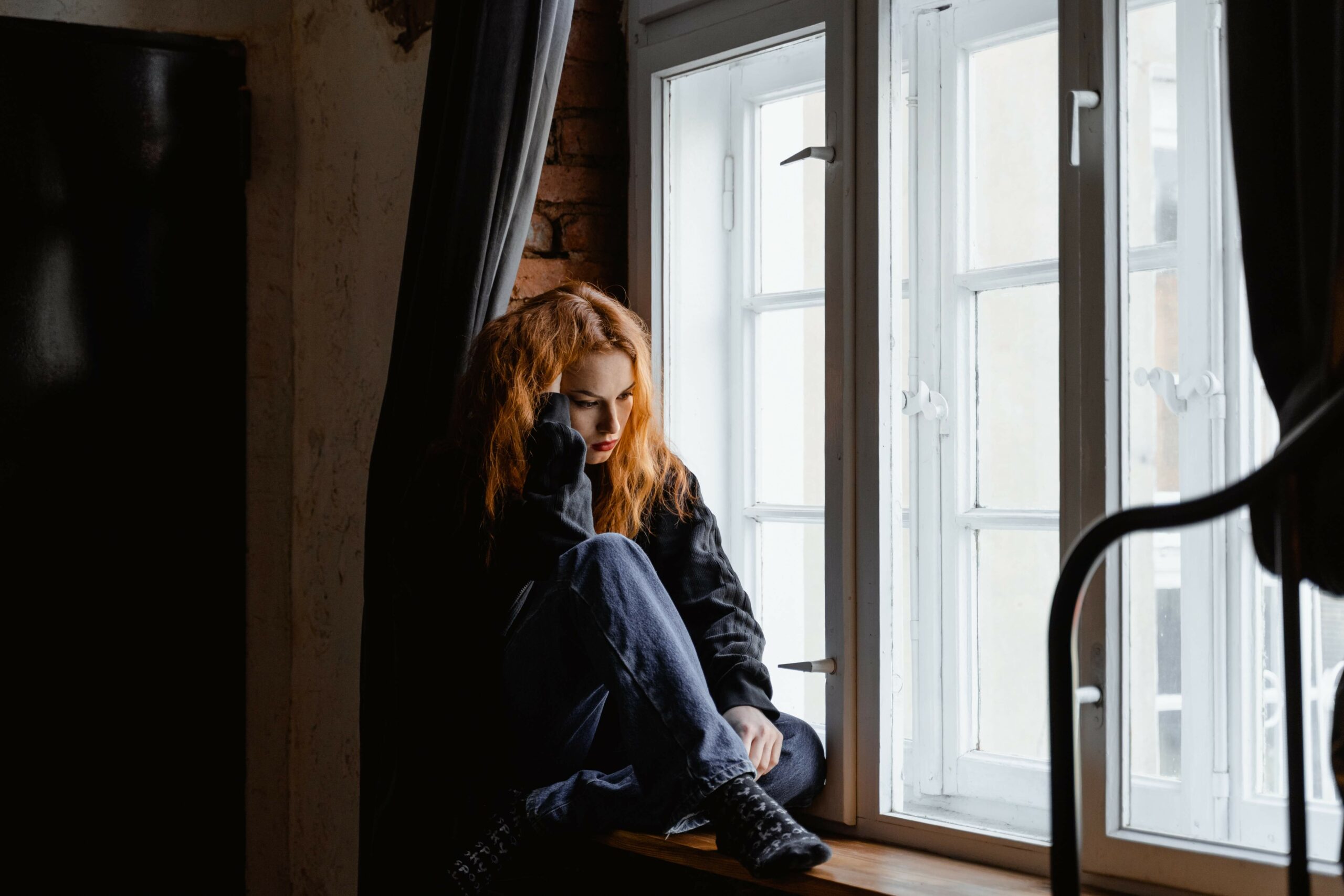 A red headed woman sits by the window dealing with the effects of PTSD. Online PTSD Treatment in Ohio, Kentucky, and New York is here to help.