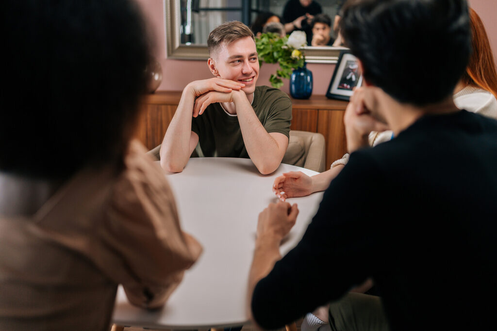 A young man sits at a table with his friends and family as he finds comfort and support with them after losing a loved one to suicide. Survivors guilt can be crippling, get the help you need in Trauma Therapy in Ohio!