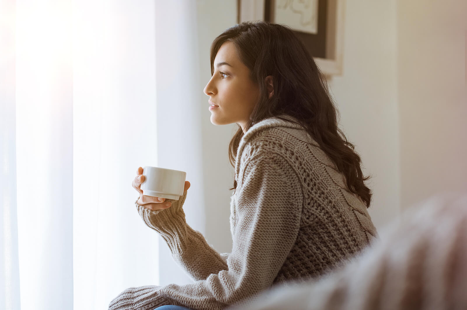 A woman sits quietly with a warm mug, looking out the window in a reflective moment after loss. This represents grief counseling in ohio, support from a grief counselor in ohio, and grief counseling in cincinnati, oh.